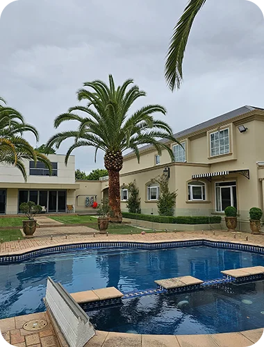 A large, luxurious, multi-story house stands behind a blue swimming pool and tall palm trees on a cloudy day.